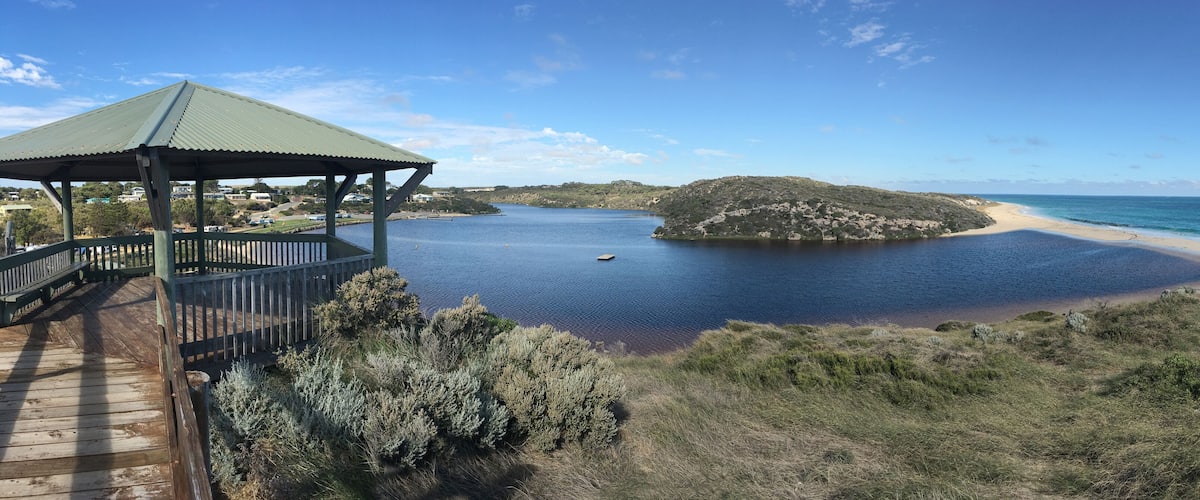 A coastal landscape with a wooden deck, a green-roofed pavilion, a sandy beach, and a small island.