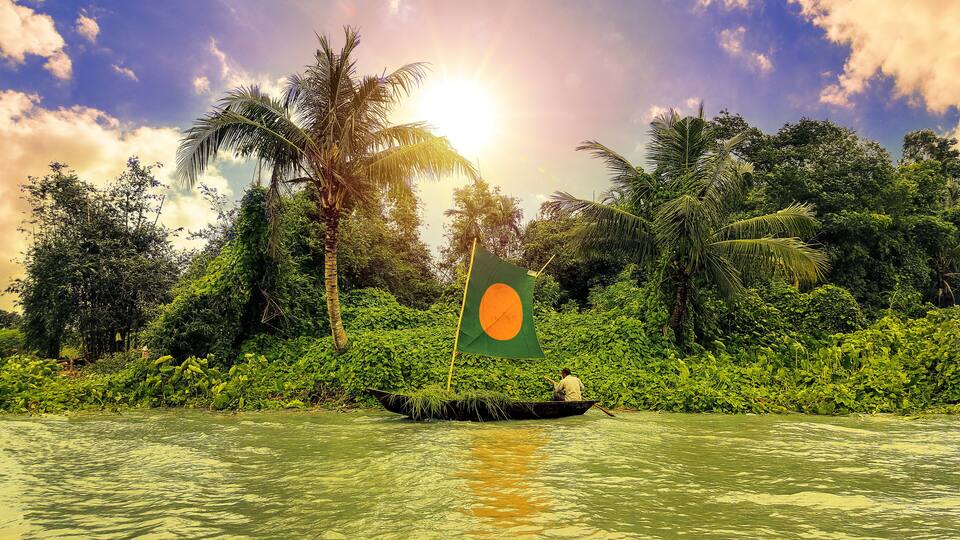 A boat with a flag on a river surrounded by dense greenery and palm trees.