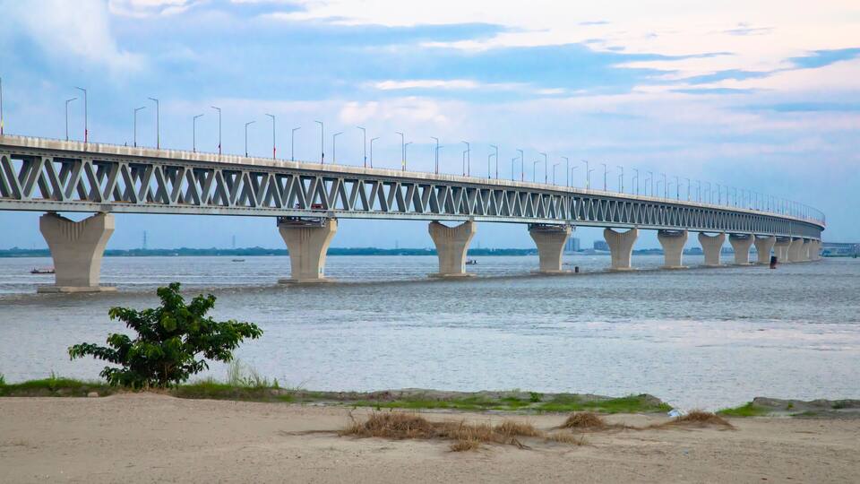 A long bridge with multiple pillars over a body of water, a sandy beach in the foreground, and a cloudy sky.