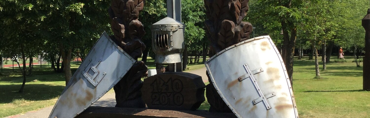 A monument with a sword and shields, labeled "ZAGGIRIS," in a park with trees and grass.