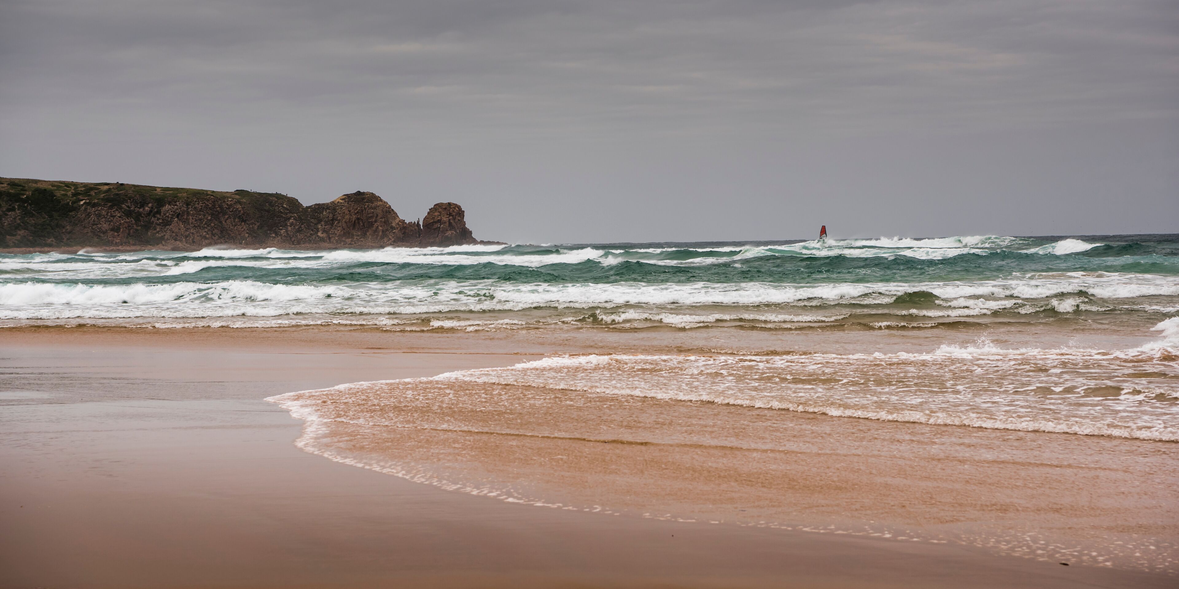 A beach with waves, a rocky cliff, and a red buoy in the distance.
