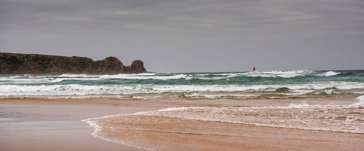 A beach with waves, a rocky cliff, and a red buoy in the distance.