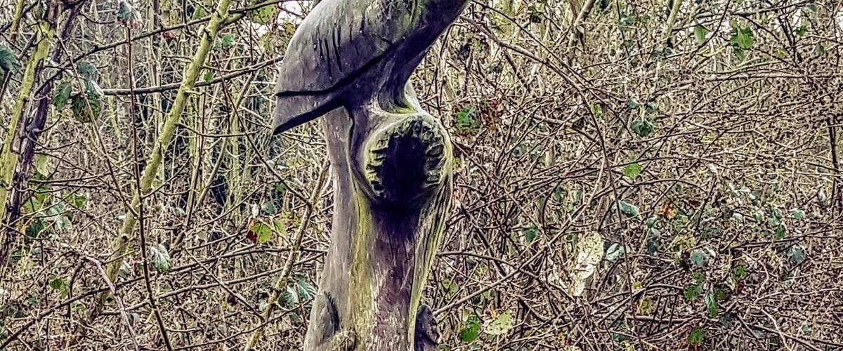 A wooden sculpture of a figure balancing a bowl on its head, set on a tree stump in a forested area.