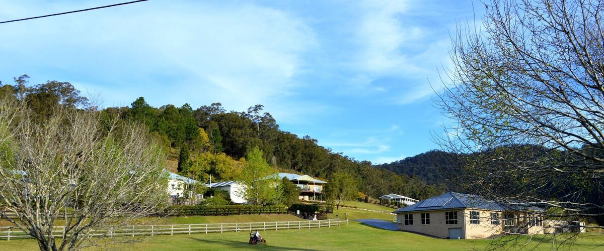 A rural landscape with a fenced field, a house with a blue roof, and a hill with trees in the background.