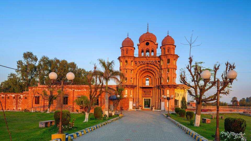 A large, ornate building with three domes and a central archway, surrounded by a paved walkway and greenery.