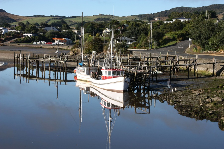 Un barco blanco amarrado a un muelle con una cabina roja, rodeado de agua tranquila y un paisaje rural con casas y colinas al fondo.