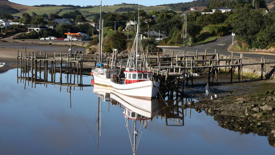 A white boat docked at a pier with a red cabin, surrounded by calm water and a rural landscape with houses and hills in the background.