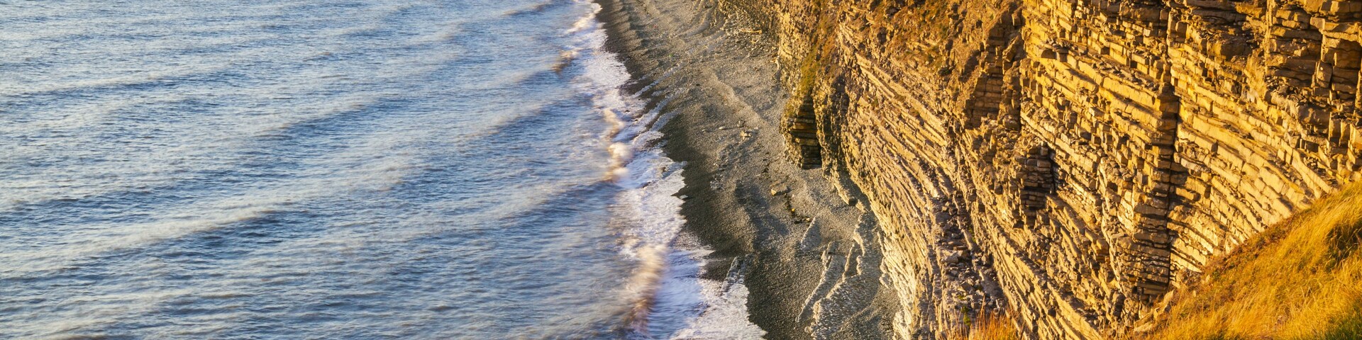 Un acantilado costero con formaciones rocosas estratificadas y una playa arenosa.