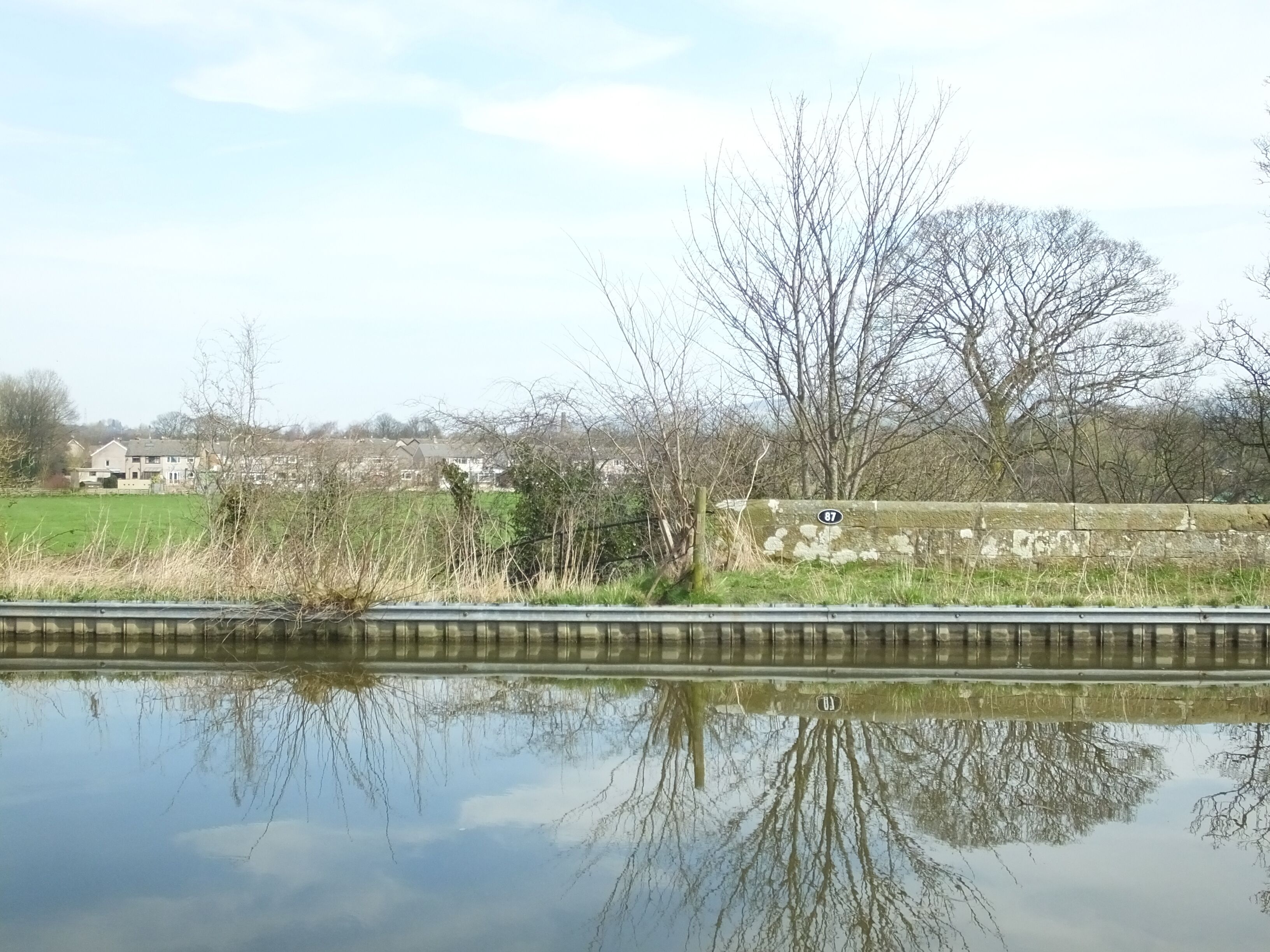 A calm river with a stone wall and a tree reflecting in the water. Houses and fields are visible in the background.