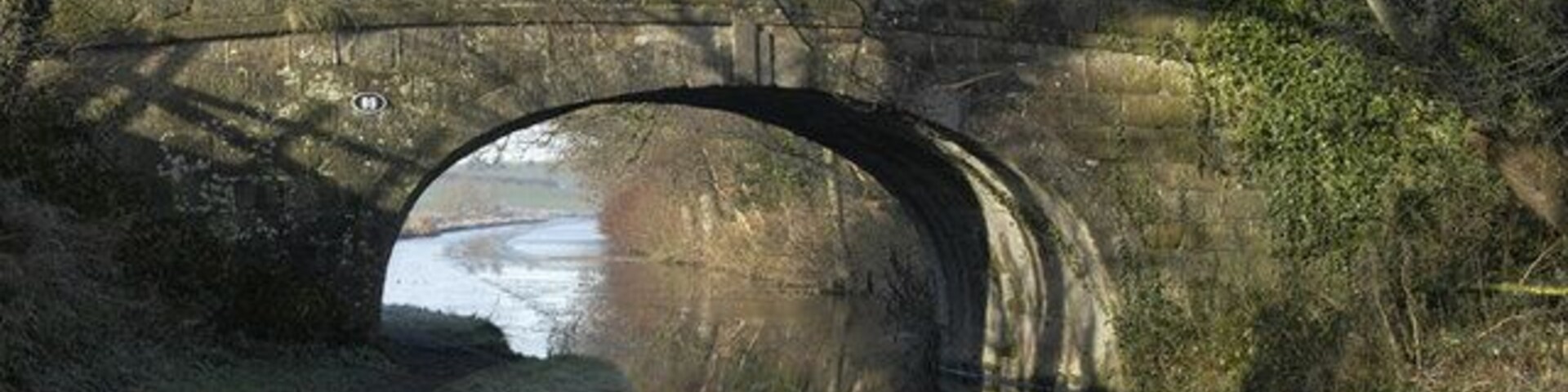 Uma ponte de pedra sobre um rio calmo, com árvores e arbustos ao redor.