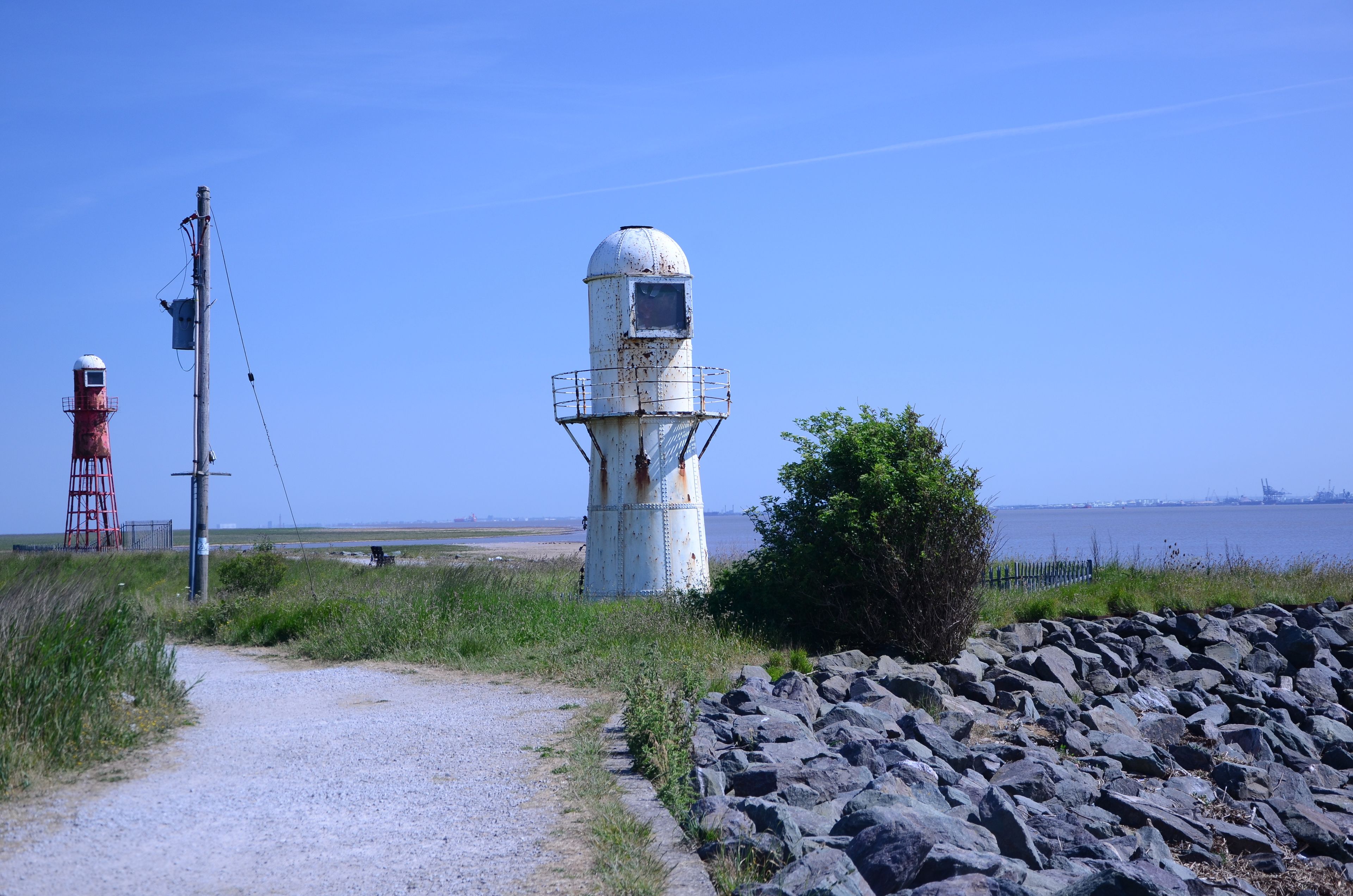 Een kustpad met een vuurtoren, rotsachtige kustlijn en een industriële haven op de achtergrond.