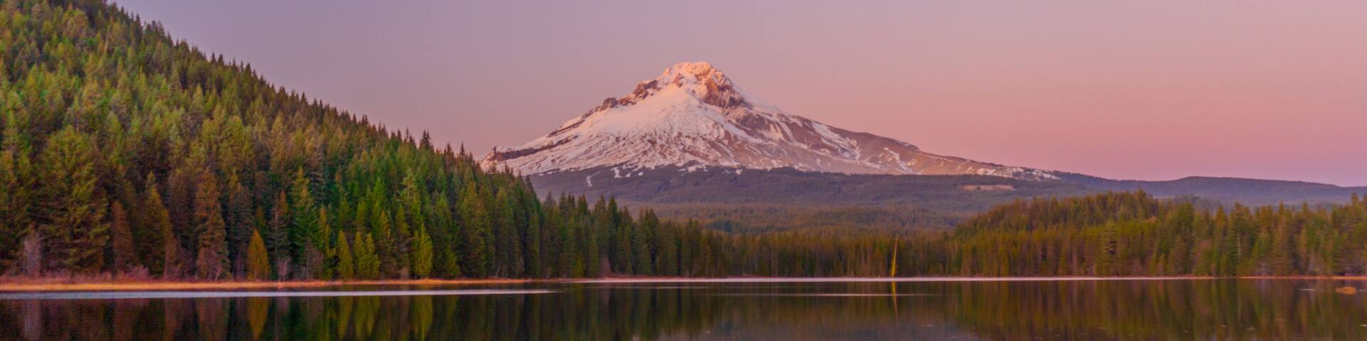 A calm lake reflecting a forest and a snow-capped mountain.