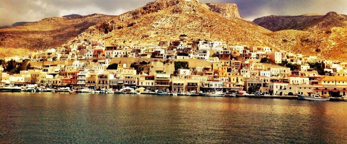 A coastal town with buildings clustered along the waterfront, a mountain in the background, and boats docked at the harbor.