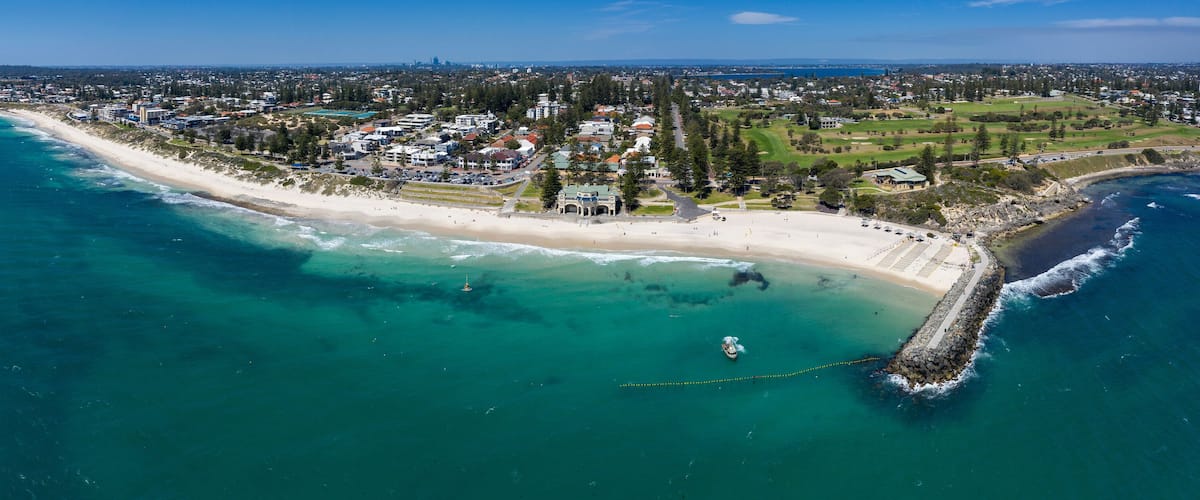 An aerial view of a coastal town with a long sandy beach, clear turquoise waters, and a mix of residential and commercial buildings.