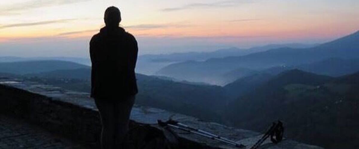 A person standing on a stone wall overlooking a valley during sunset.