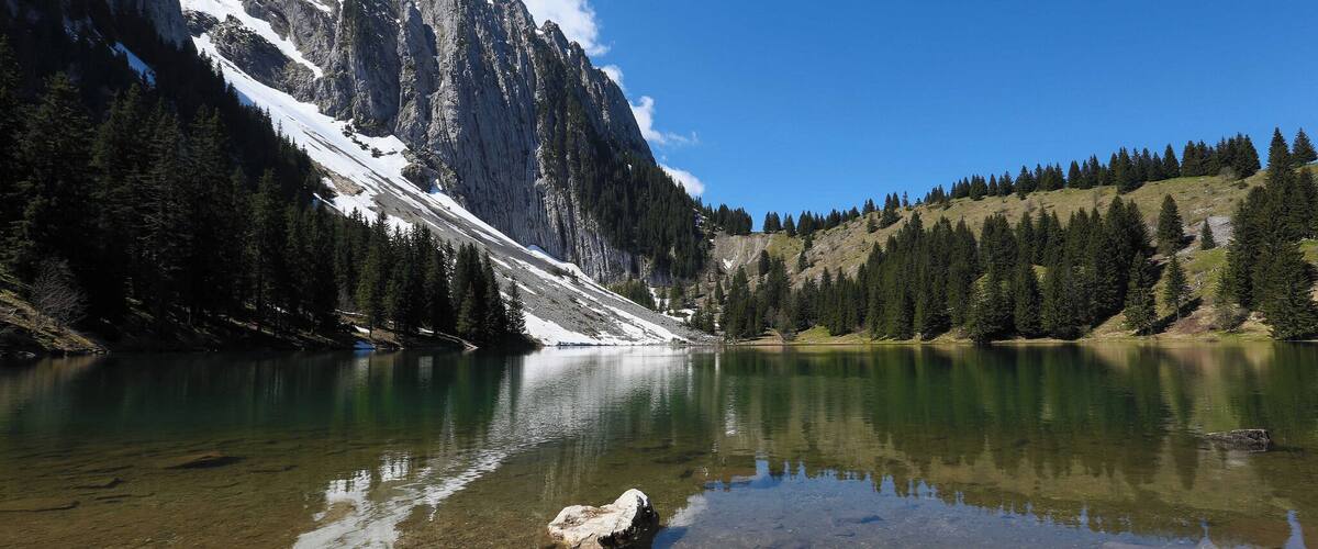 A mountain lake with clear water, surrounded by snow-capped peaks and dense forests.