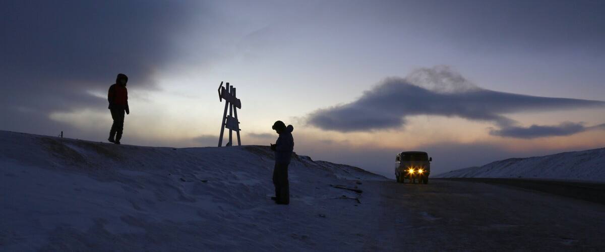 A snowy landscape with three people standing on a hill, a vehicle with headlights on, and a cloudy sky.