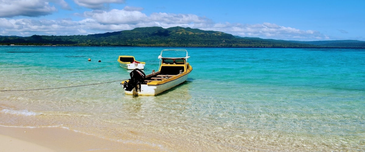 A small boat tied to a sandy beach with clear turquoise water and a mountain in the background.