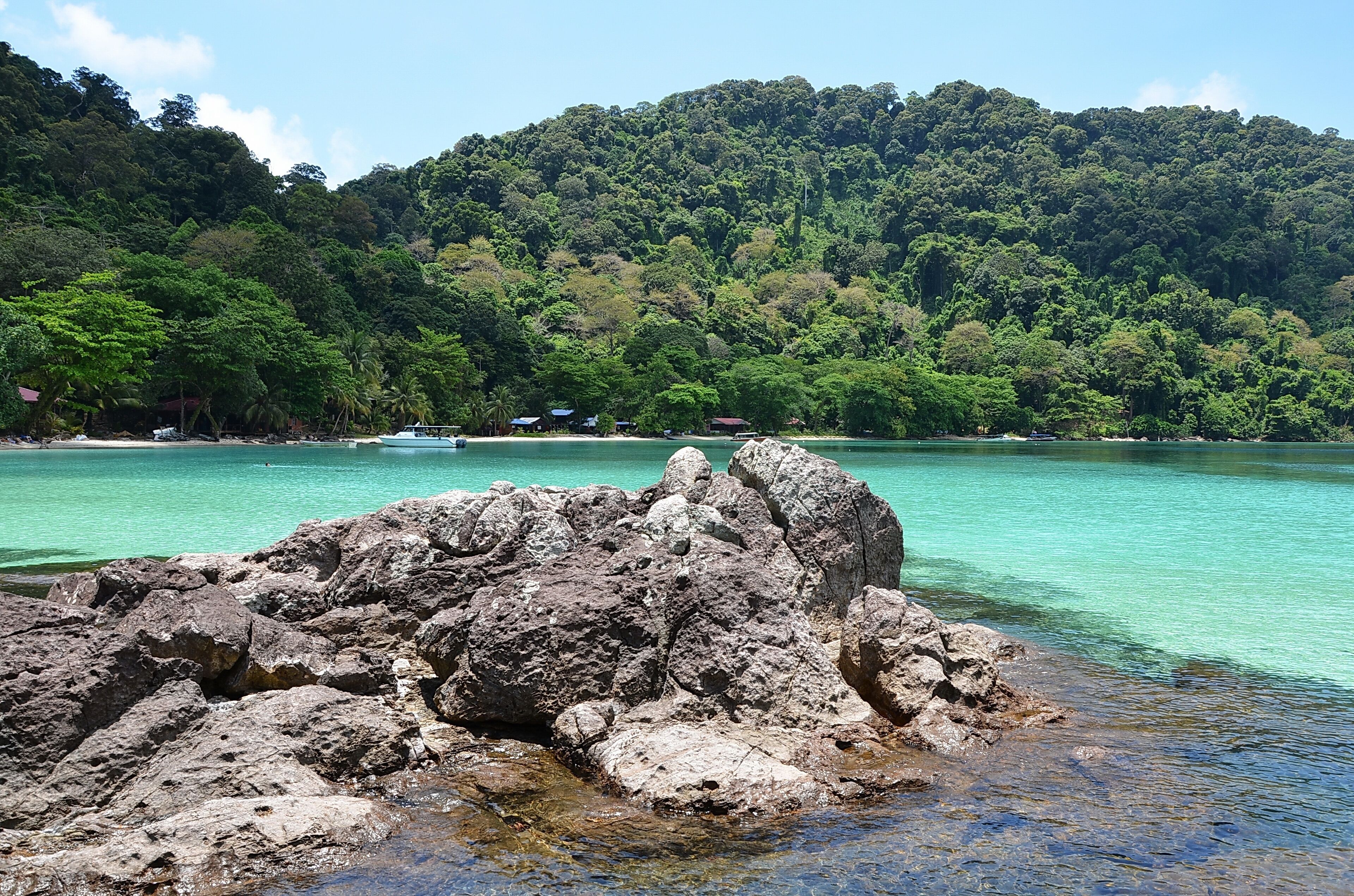 A rocky shoreline with clear turquoise water, a forested hill in the background, and a few small buildings near the water's edge.