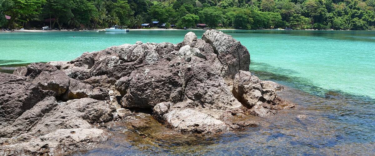 A rocky shoreline with clear turquoise water, a forested hill in the background, and a few small buildings near the water's edge.