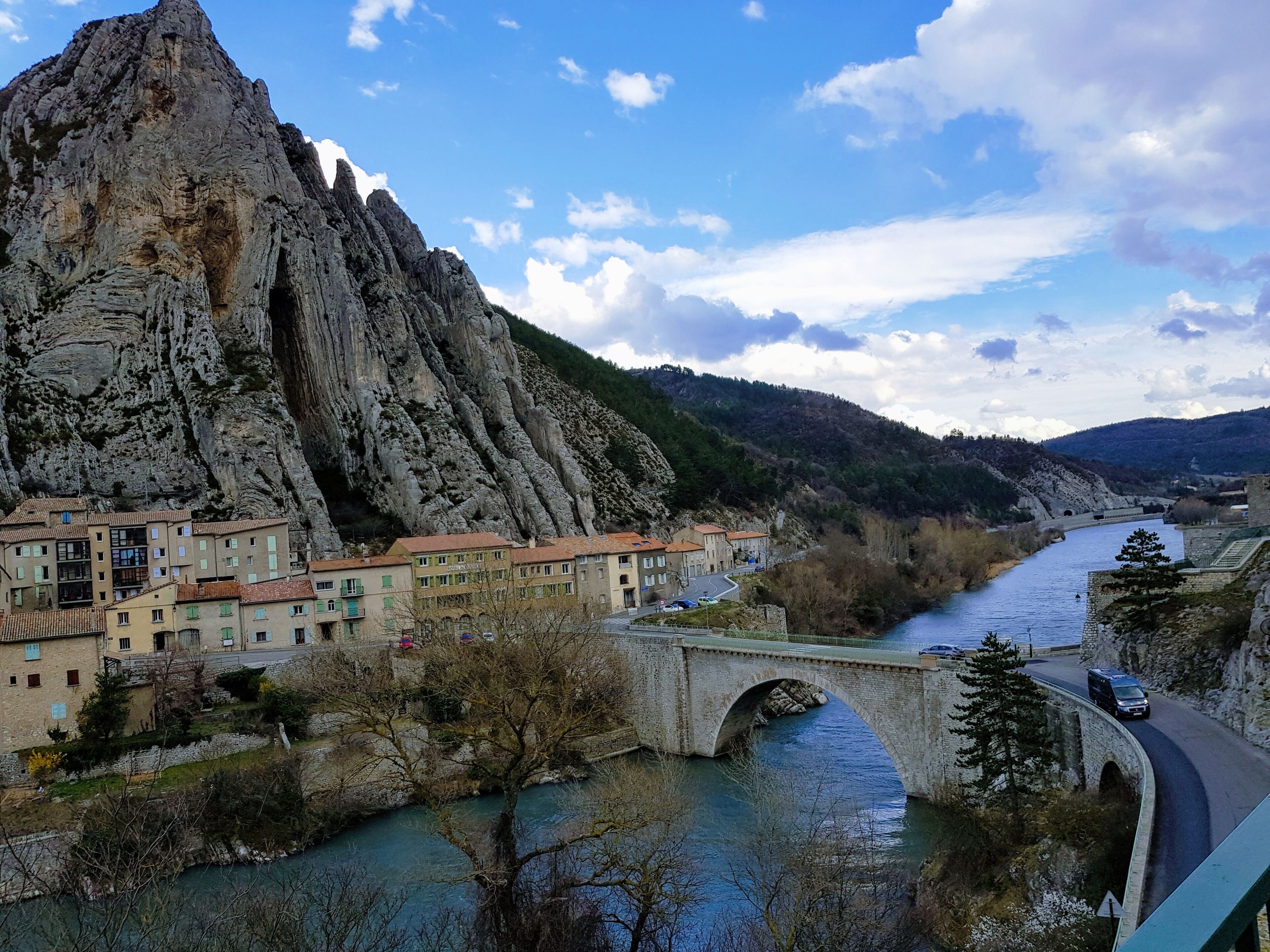 A river flowing through a town with a bridge and a large rocky mountain in the background.