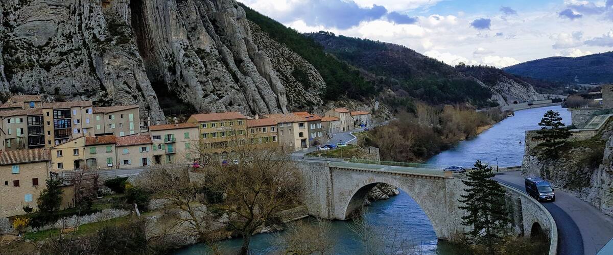 A river flowing through a town with a bridge and a large rocky mountain in the background.