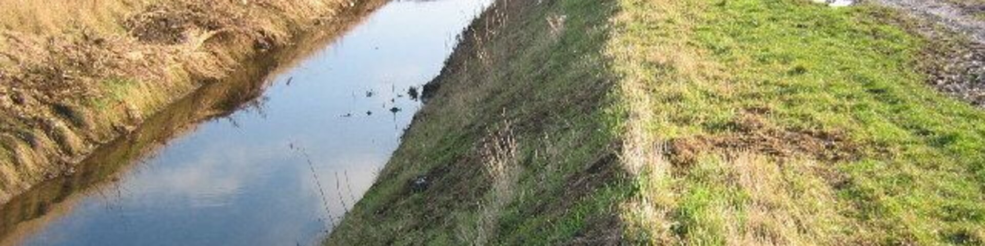 Um canal de água estreito, com margens gramadas e campos de ambos os lados.