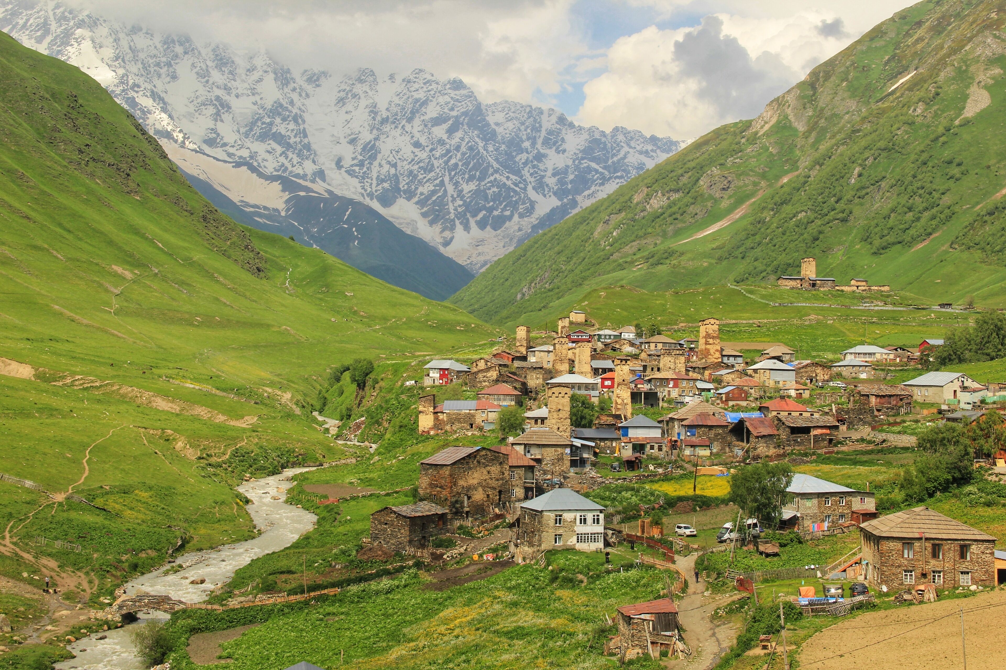 A village nestled in a valley with a river, surrounded by green hills and snow-capped mountains in the background.
