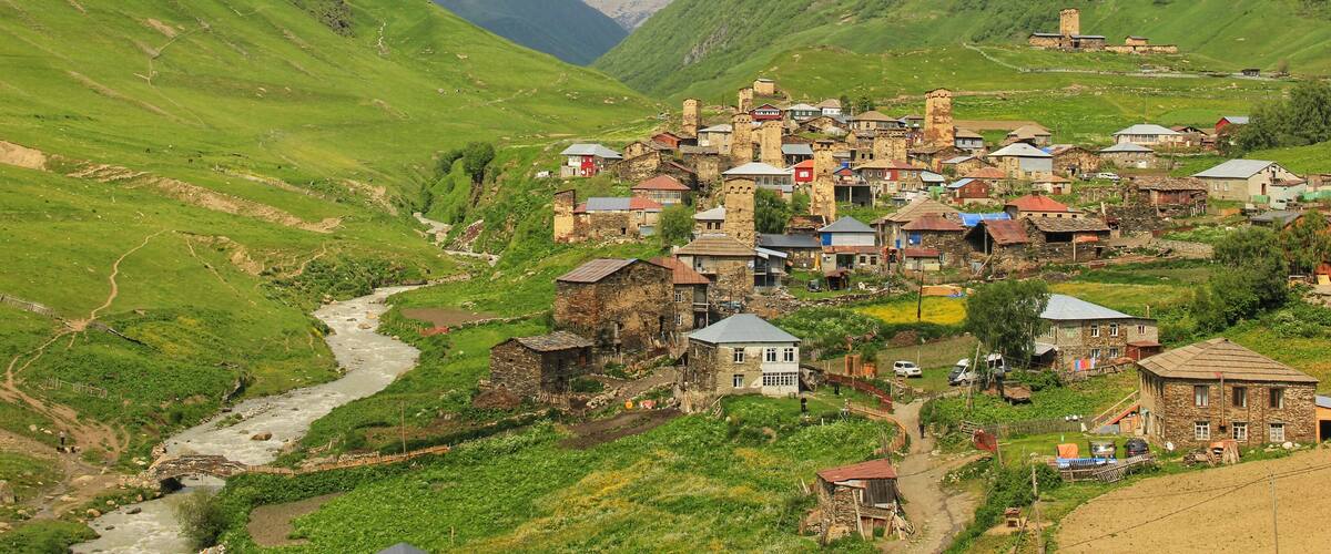 A village nestled in a valley with a river, surrounded by green hills and snow-capped mountains in the background.