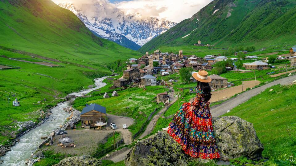 A mountainous village with a river, houses, and a person in a colorful dress.