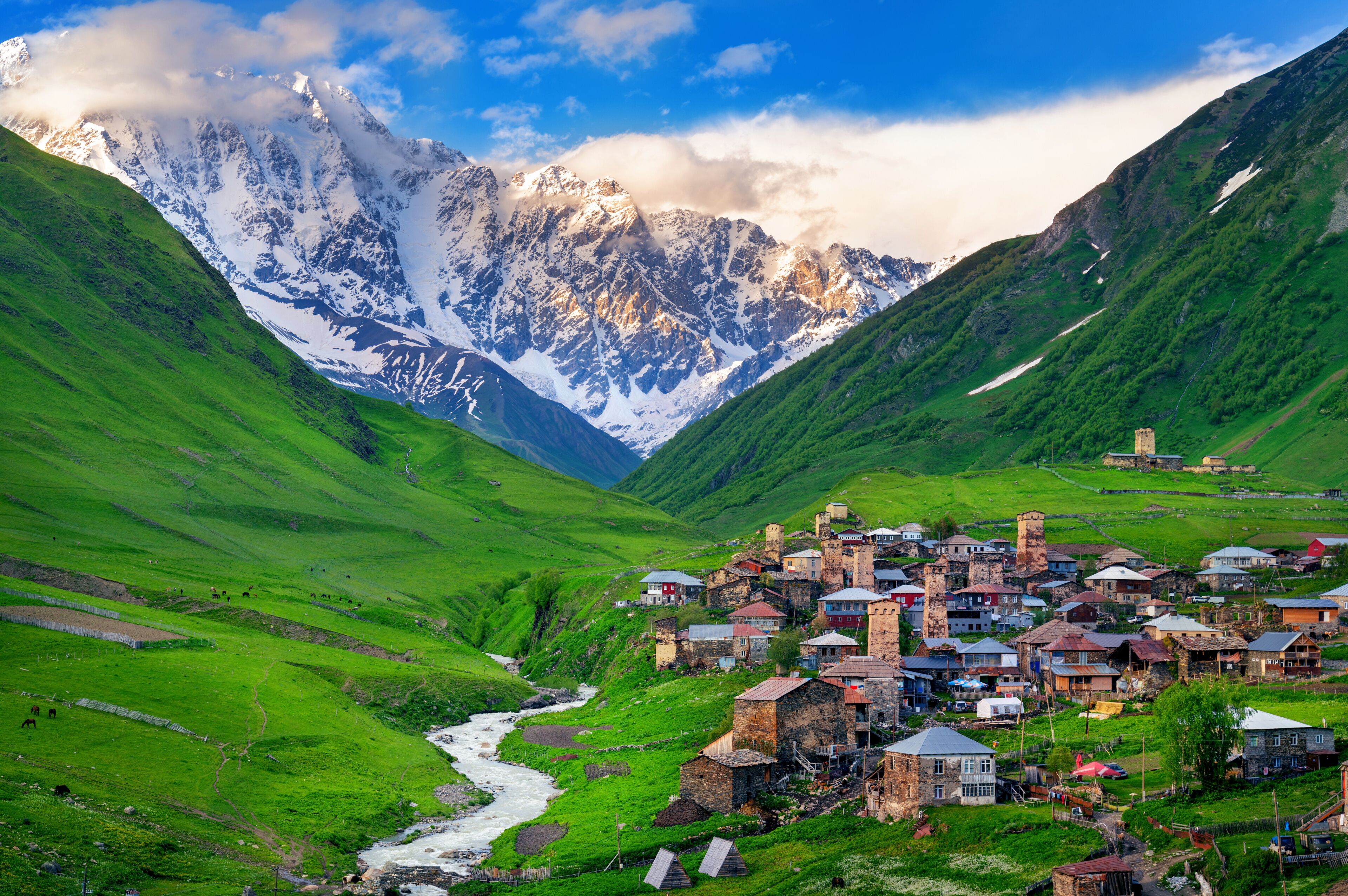 A village nestled in a valley with a river, surrounded by green hills and snow-capped mountains.