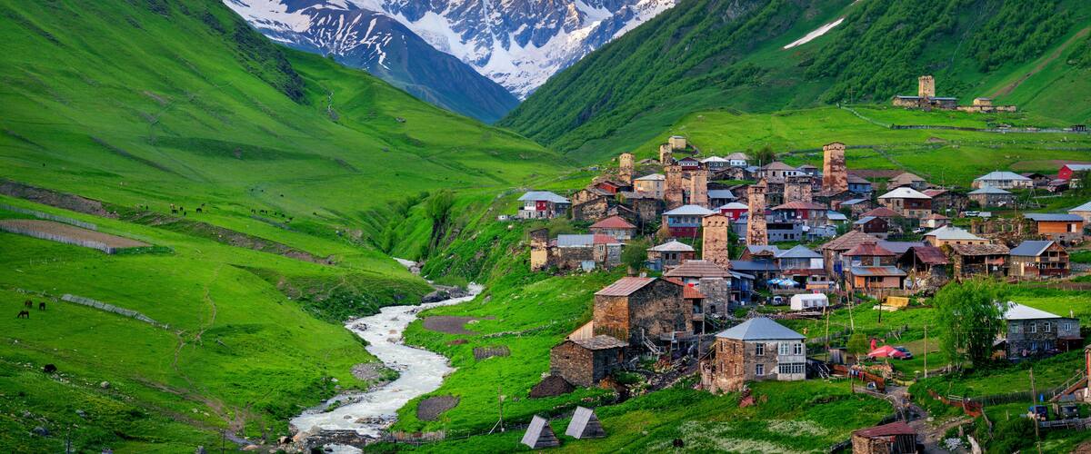 A village nestled in a valley with a river, surrounded by green hills and snow-capped mountains.