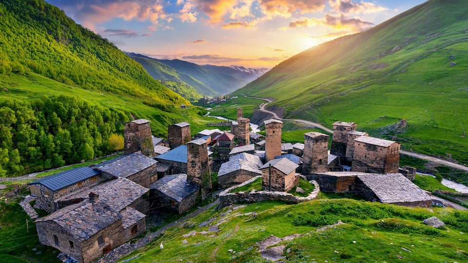 A medieval village with stone buildings and towers, surrounded by lush green hills and a valley.