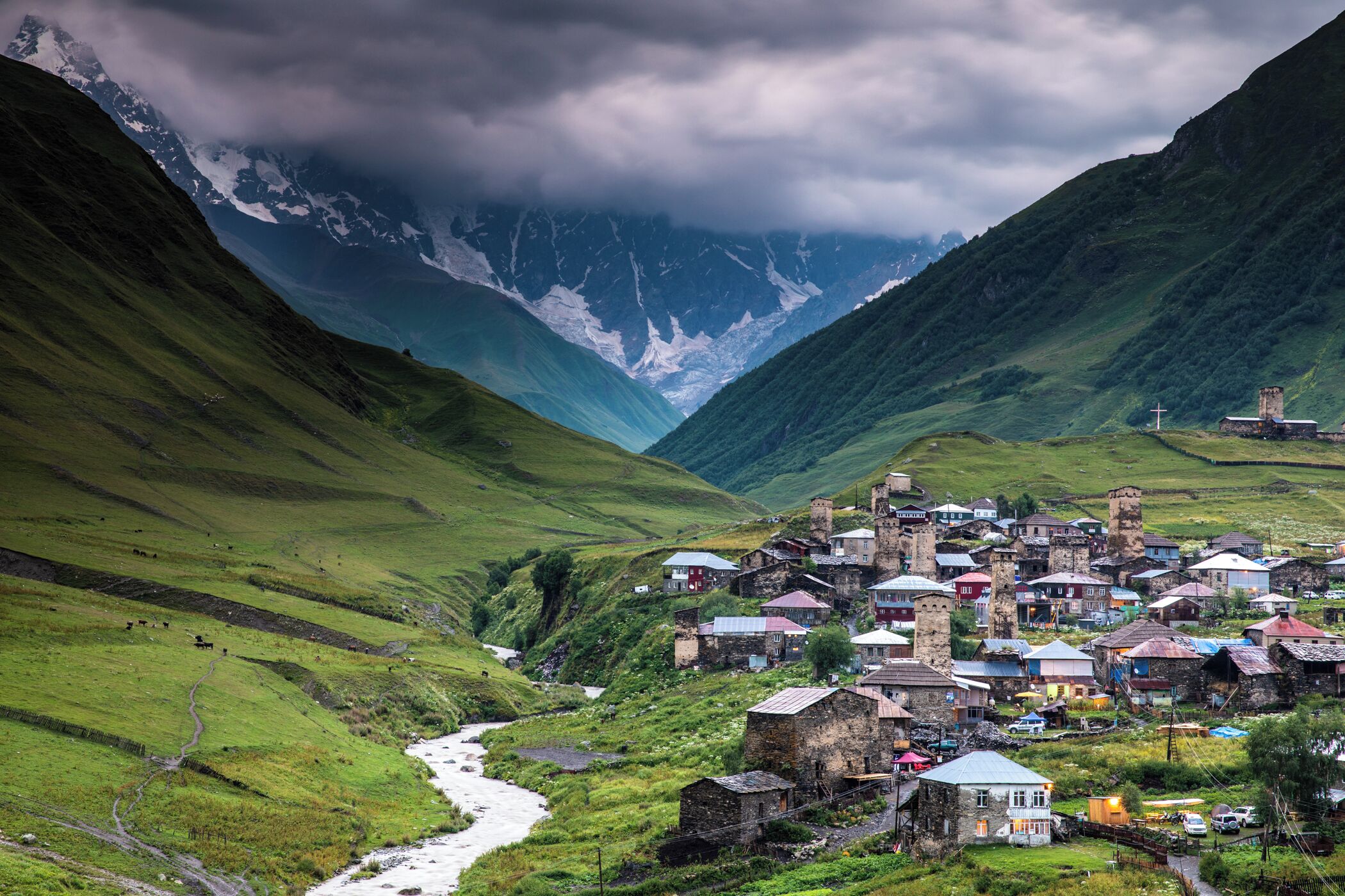 A village nestled in a valley with a river, surrounded by mountains and a cloudy sky.
