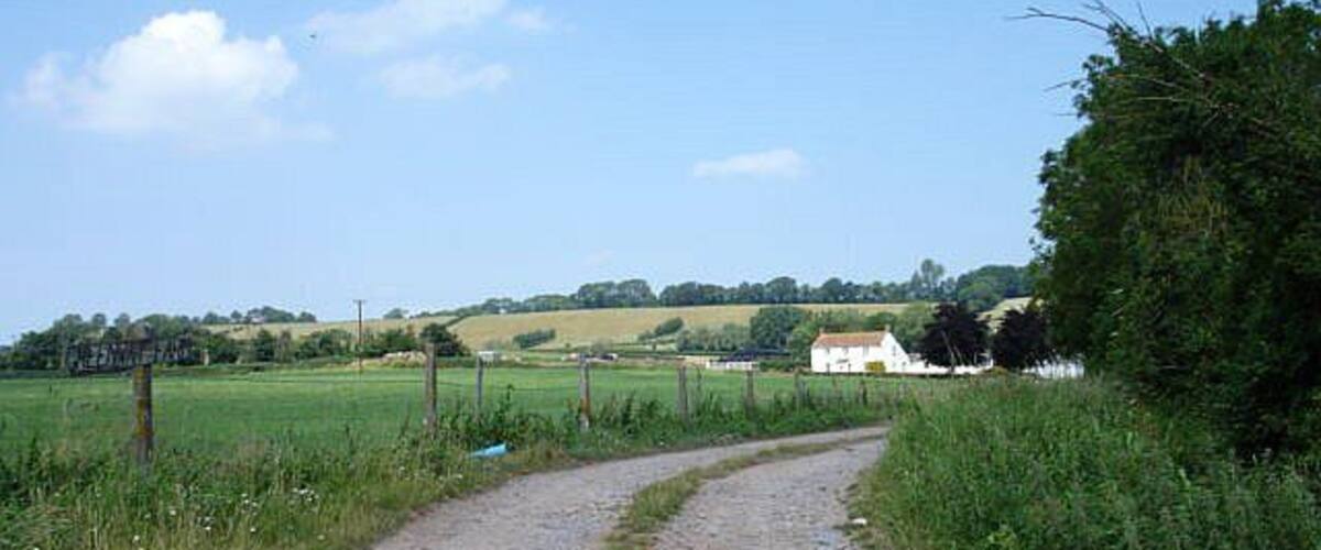 A gravel path curving through a rural landscape with fields and a white house in the distance.