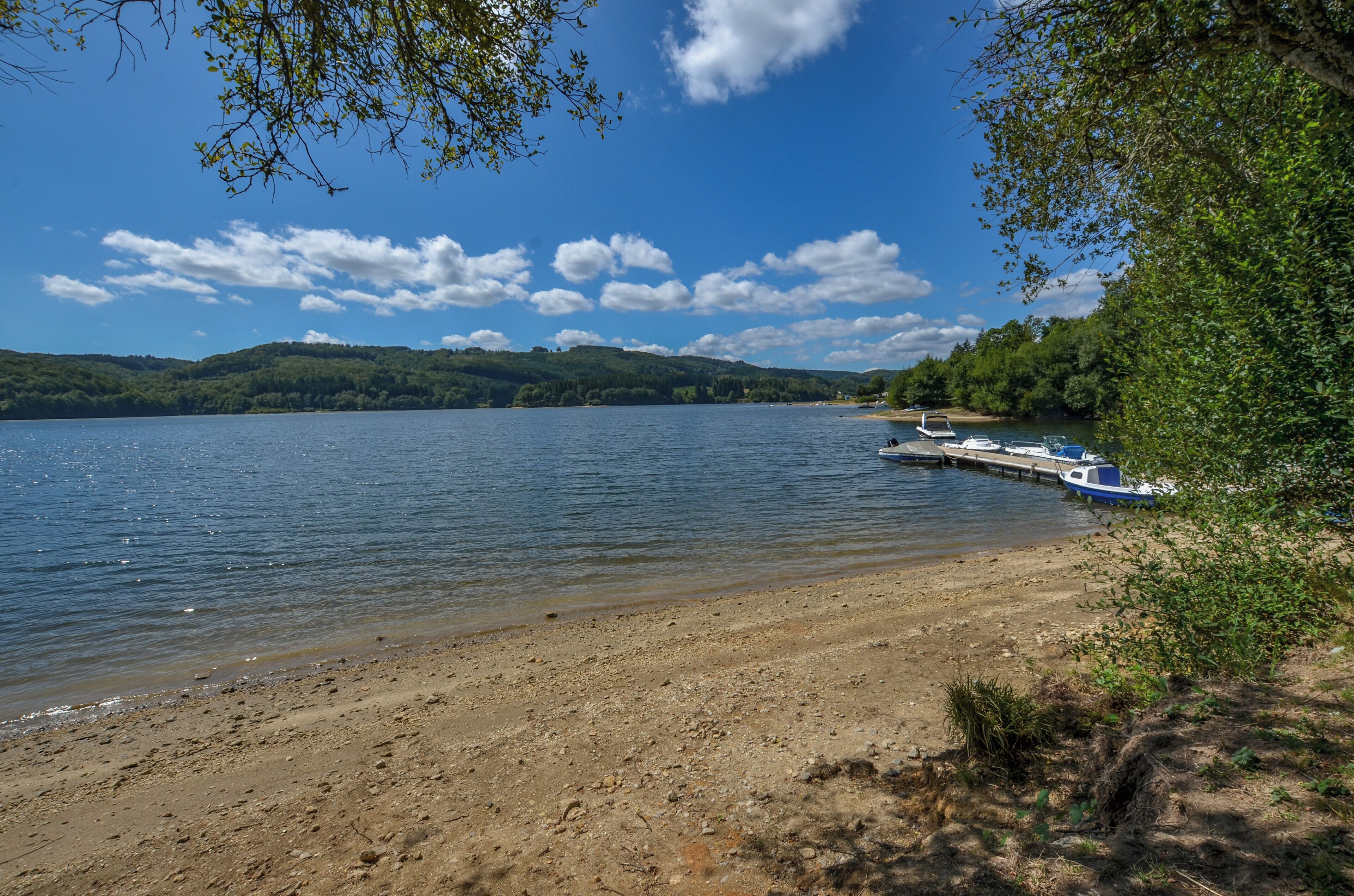 A lake with a sandy shore, a dock with boats, and green hills in the background.