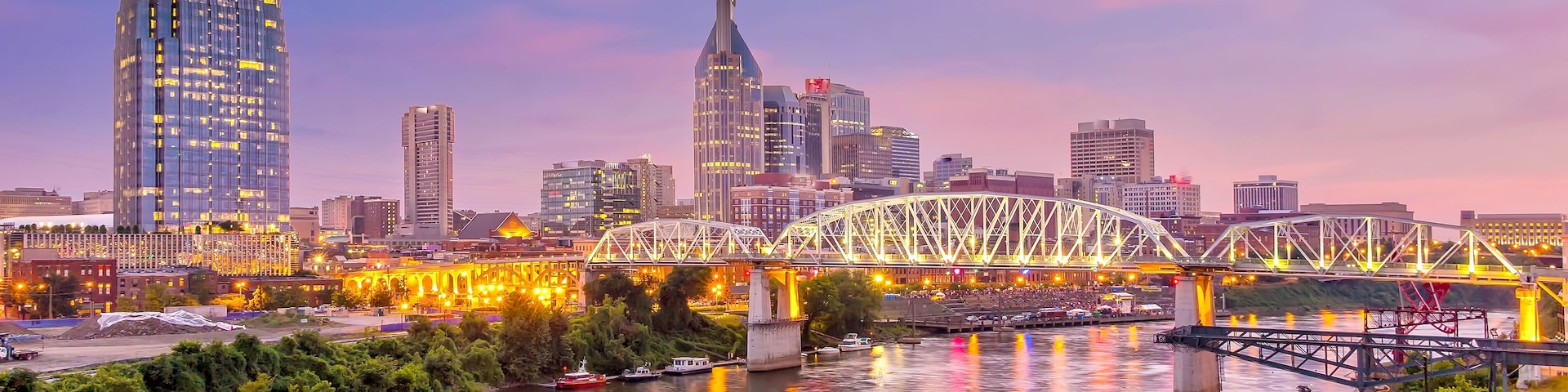A river with a brightly lit boat, a bridge, and a city skyline with modern buildings.