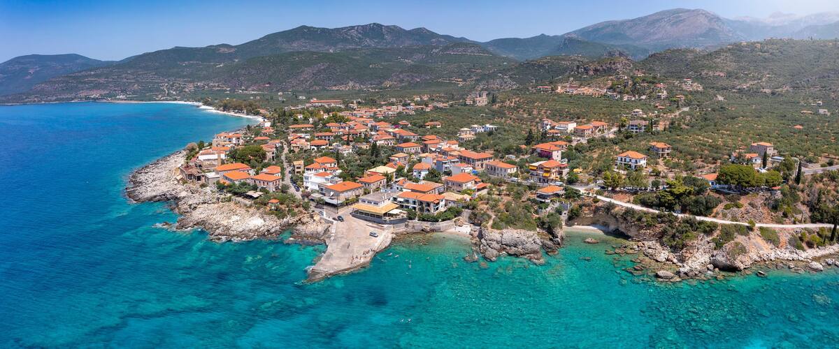 A coastal town with clear turquoise waters, red-roofed buildings, and mountainous terrain in the background.