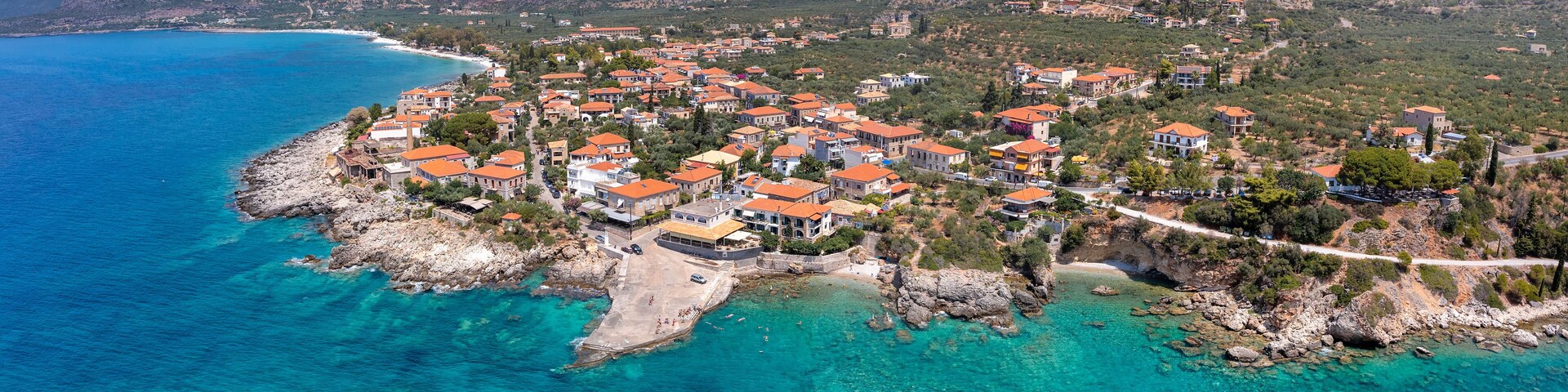 A coastal town with clear turquoise waters, red-roofed buildings, and mountainous terrain in the background.