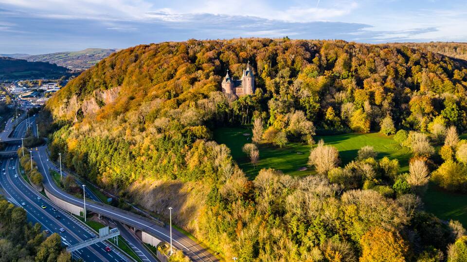 A castle surrounded by autumn foliage, a highway, and a small body of water.