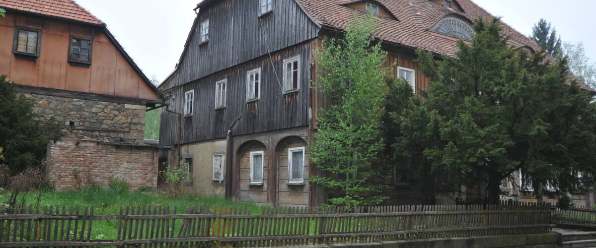 A historic building with a wooden facade and a tiled roof, surrounded by trees and a stone wall.