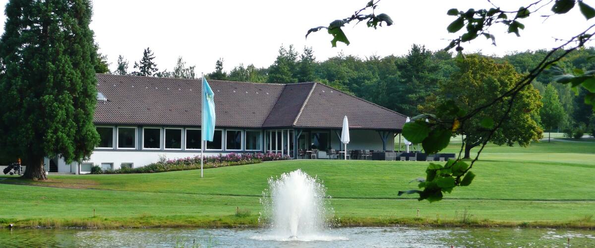 A golf course with a pond, a building with a flag, and trees in the background.