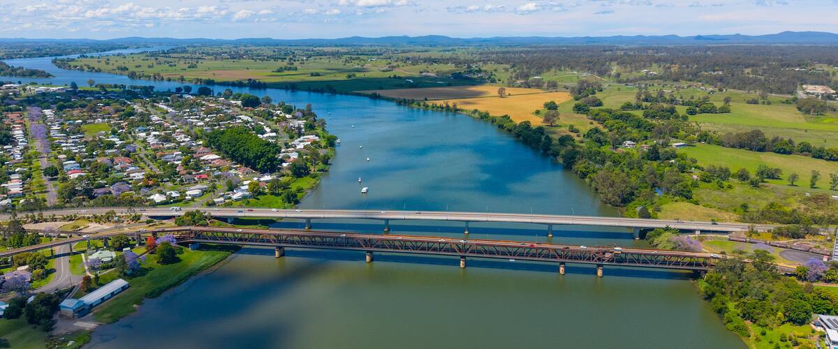 A river flowing through a town with a bridge, surrounded by green fields and distant mountains.