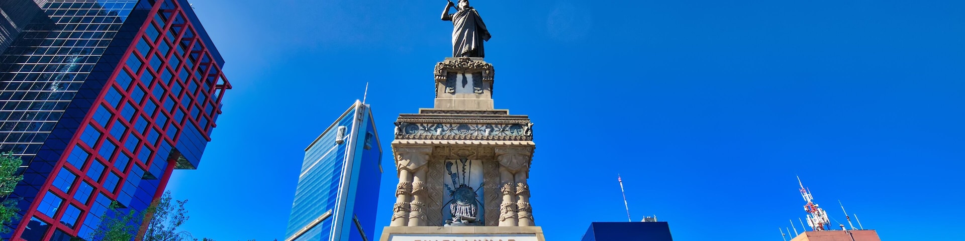 Un monumento con una estatua e inscripciones, rodeado de edificios modernos y árboles.