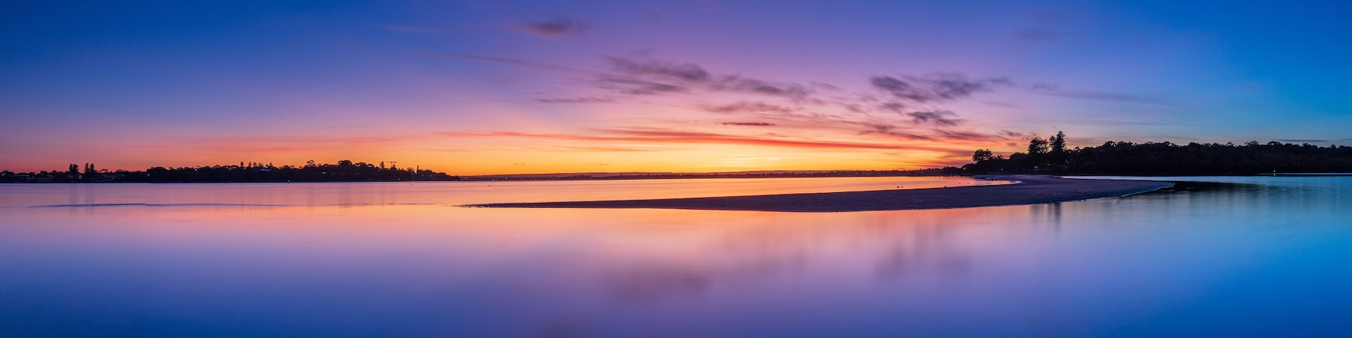 Un lago tranquilo al atardecer, con el reflejo del cielo, una orilla distante y un horizonte despejado.