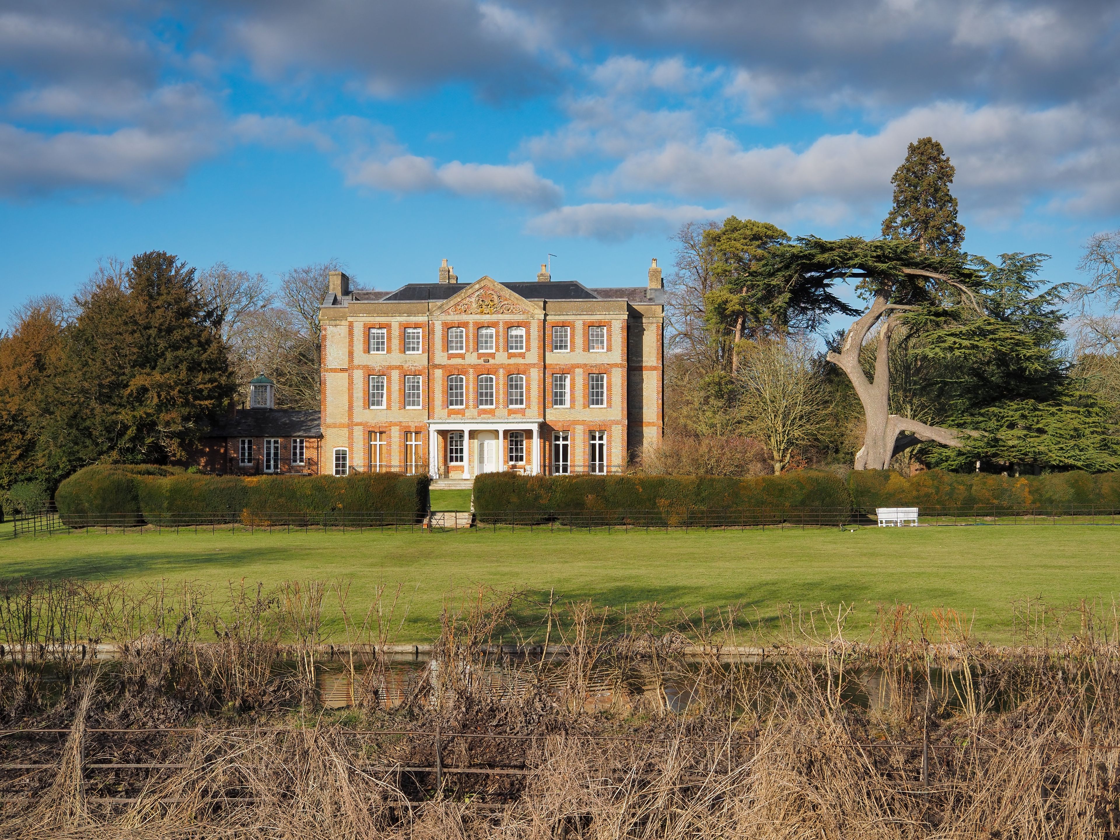 The perfectly symmetrical Baroque style Ardington House, commissioned by Edward Clarke in 1720, framed by interesting trees and under blue sky and clouds, Oxfordshire, UK