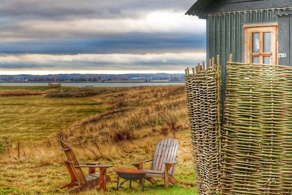 Een rustieke houten constructie met een raam, omgeven door een vlechtwerk hek, en twee houten stoelen en een tafel in een grasveld onder een bewolkte lucht.