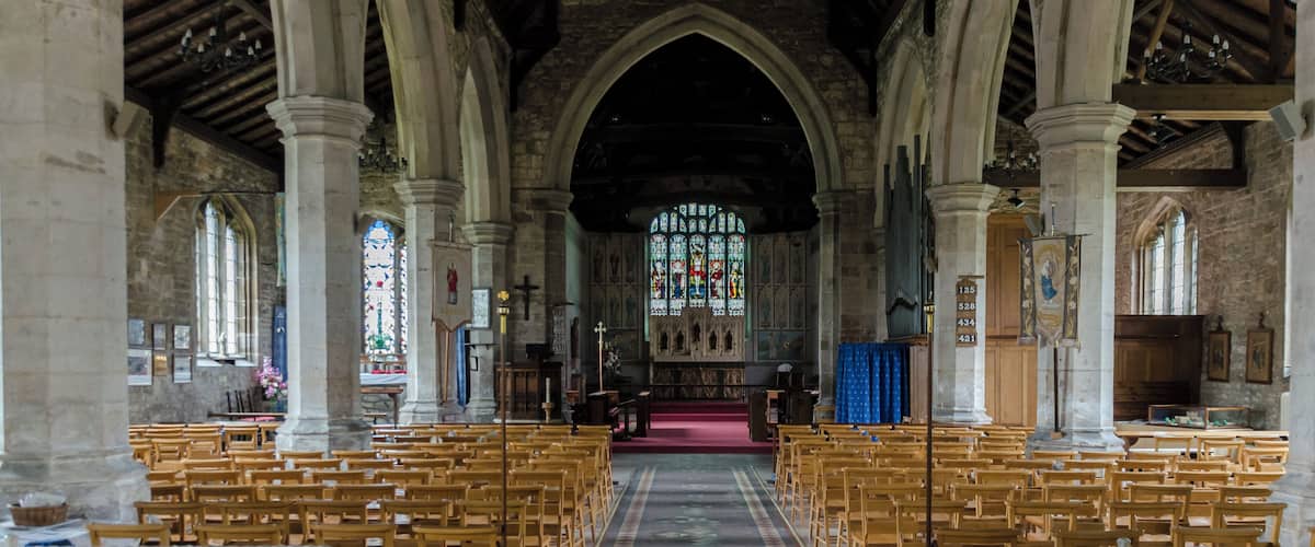 An interior of a church with wooden pews, a central aisle, stained glass windows, and stone columns.