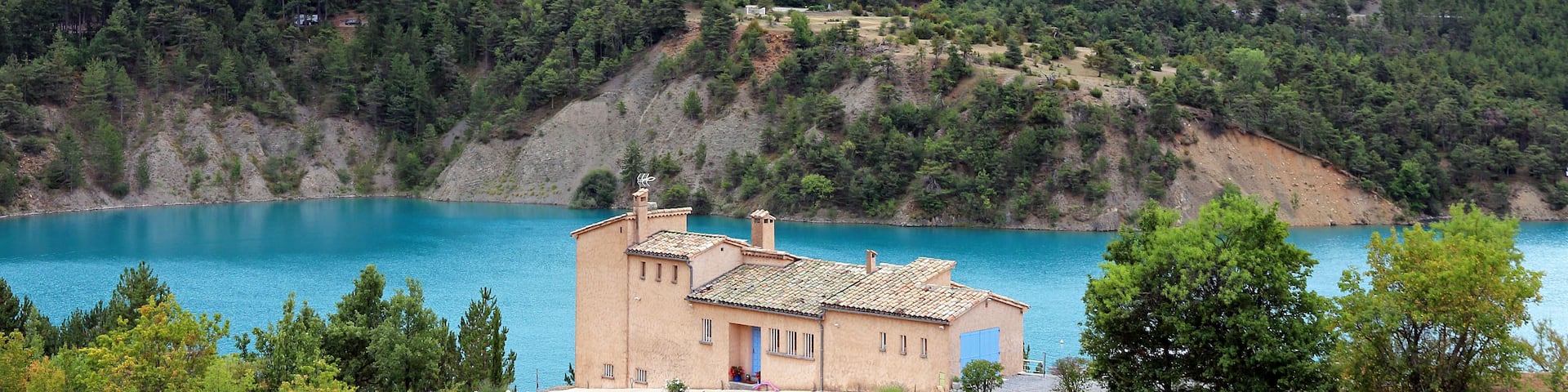 A house by a turquoise lake surrounded by forested hills.