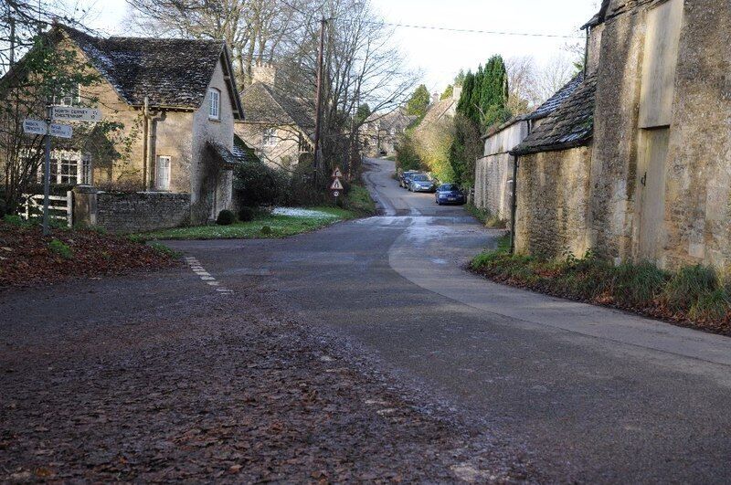 A narrow rural road flanked by stone buildings, a signpost, and a pedestrian crossing.